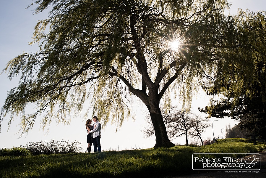 A couple stands under a large tree in the sunshine during their outdoor engagment portrait session by Seattle engagement photographer Rebecca Ellison