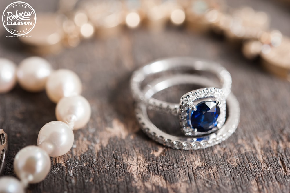 Bride's rings and pearl necklace sit on a rough wooden floor photographed by Rebecca Ellison photography
