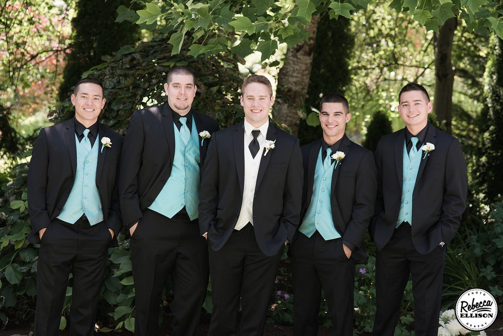 Groomsmen stand in front of a group of trees during outdoor wedding portraits featuring black suits with blue vests and ties photographed by Seattle wedding photographer Rebecca Ellison
