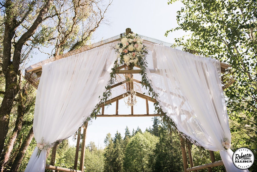 Wooden wedding arbor decorated with billowing white curtains, flowers, leaves and a crystal chandelier at an outdoor garden wedding photographed by Rebecca Ellison Photography