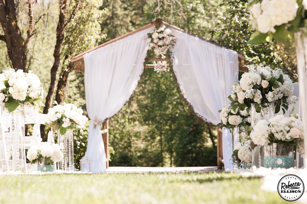 Wedding details at an outdoor garden wedding featuring a wooden arbor, white curtains and a flower lined wedding aisle photographed by Rebecca Ellison Photography