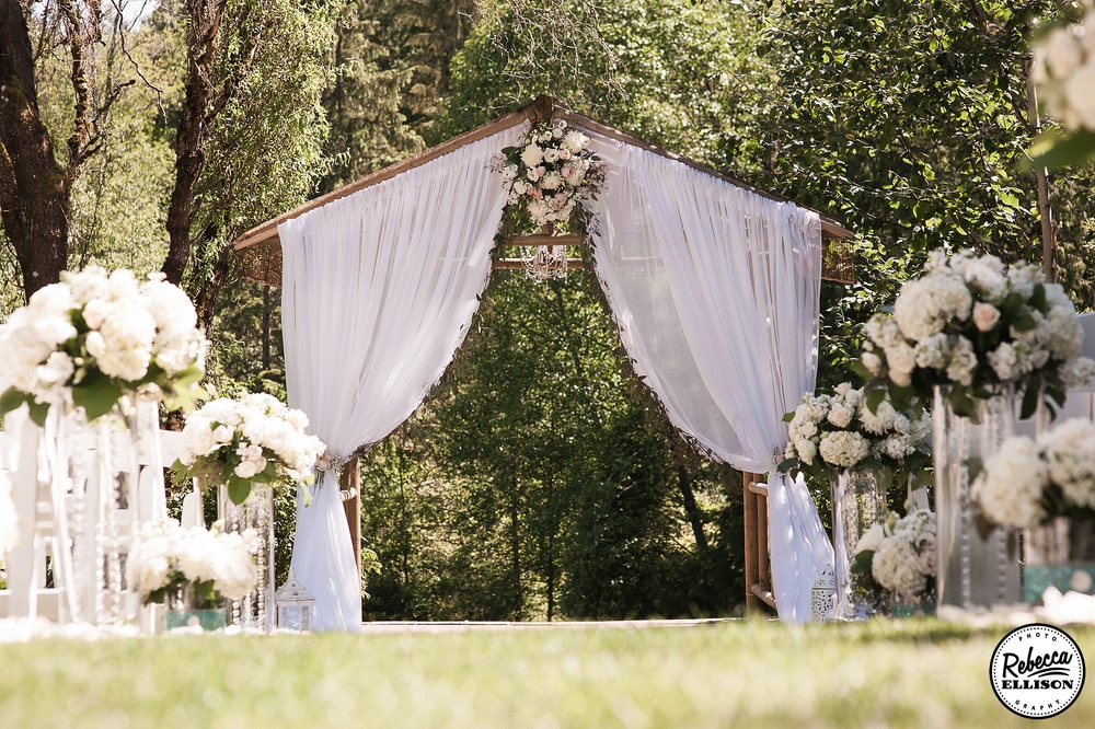 Outdoor garden wedding featuring a flower lined aisle and a wooden arbor decorated with flowers and white curtains photographed by Rebecca Ellison Photography