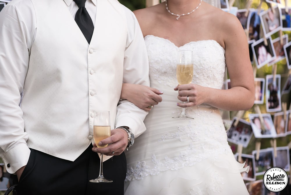 Bride and groom hold their champagne flutes for toasts in front of a photo display photographed by Rebecca Ellison Photography