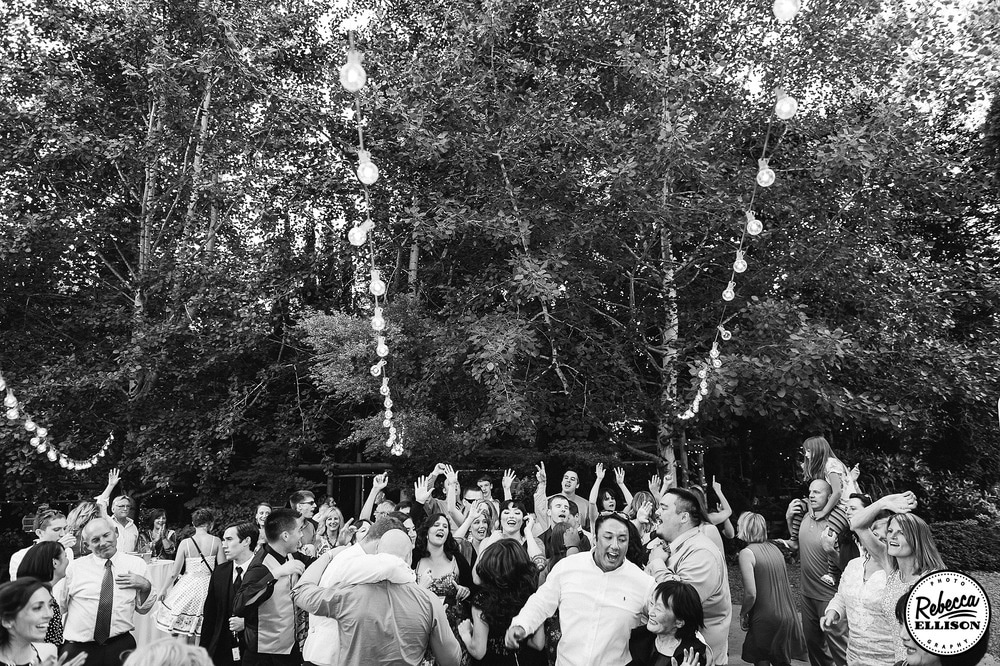 Wedding guests dance at an outdoor wedding reception featuring a forest of trees and lightbulb strings photographed by Rebecca Ellison Photography