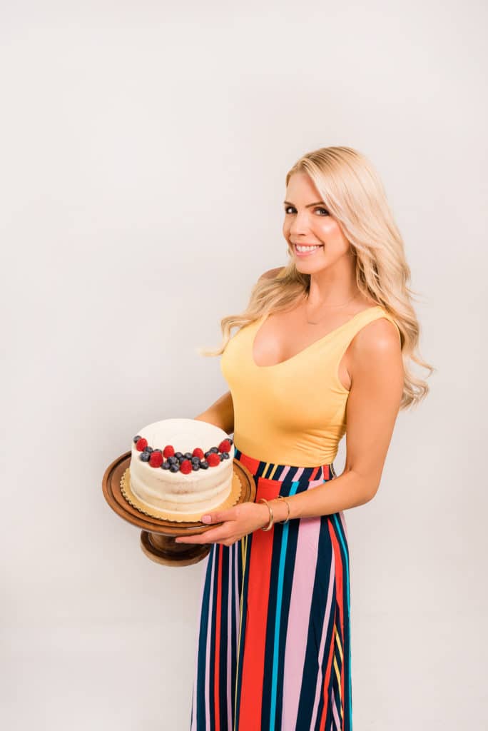 Sarah Jio wearing a yellow top and colorful striped skirt while holding a cake on a cake stand